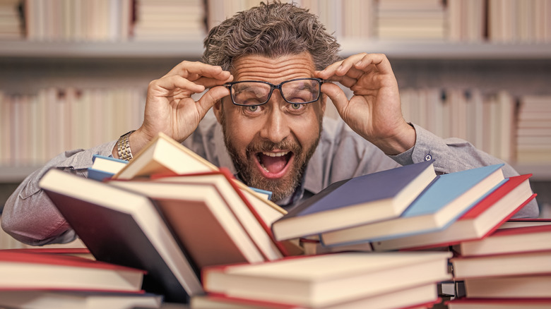 A teacher stands behind a stack of books in a library lifting his glasses off his face