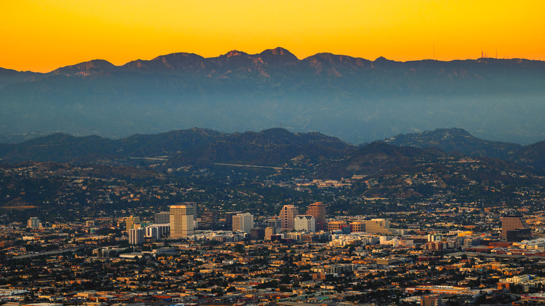 A cityscape is seen in the foreground below a mountain range and an orange sky