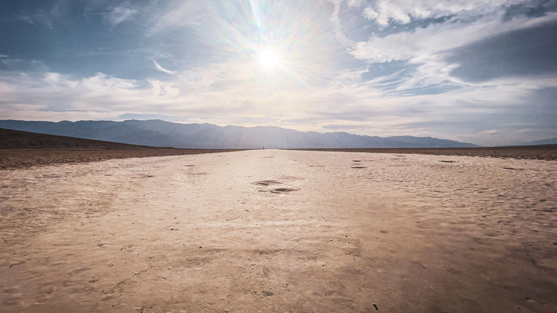 The barren desert landscape of Death Valley is seen below a cloudy sky with a bright sun shining through