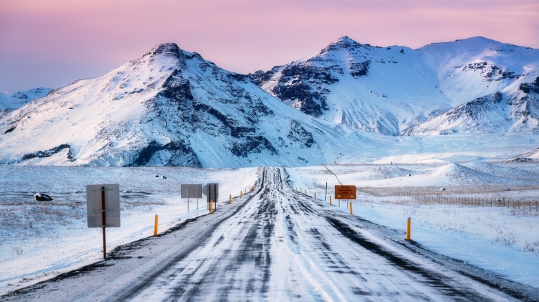 Vehicle tracks on winter road with snow-covered mountains and sunset in the background