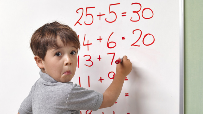 Young boy pulling face writing math problems on whiteboard with red marker