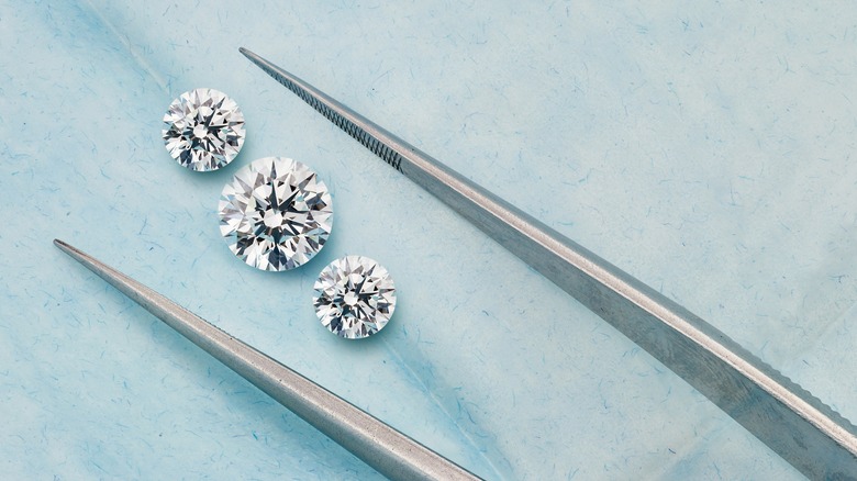 Three lab-grown diamonds sit on a surface as a pair of tweezers approaches