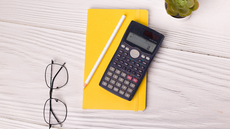 Scientific calculator on a yellow notebook with a white pencil, glasses, and a table plant