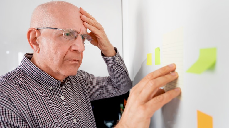 An elderly man is shown holding his head and looking at post-it notes on the wall