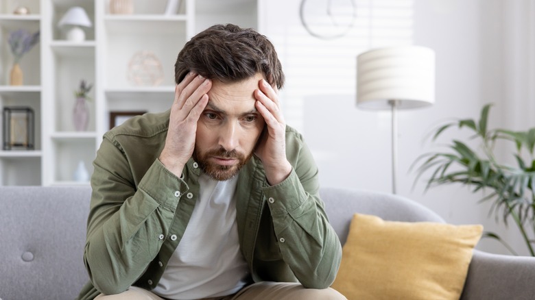 A man sits on a couch with his head in his hands looking concerned