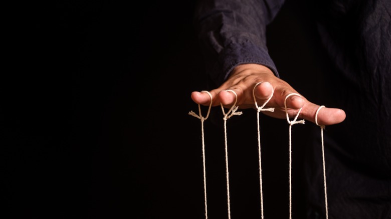 Puppet strings are shown dangling from the fingers of a hand in close-up