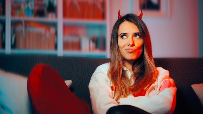 A woman sits on a couch wearing devil horns with a scheming look on her face