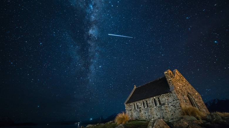 A long-exposure photo at night with a church in the foreground and a streak of light surrounding by countless stars in the sky above