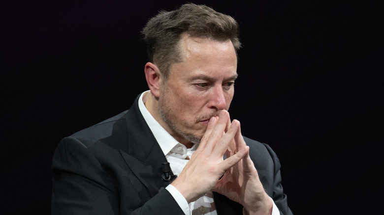 Elon Musk sits against a black background during an event looking concerned