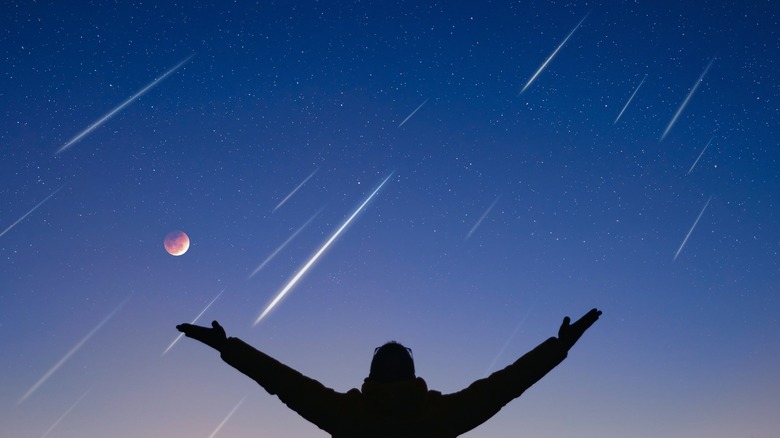 Silhouette of man with arms raised in a V, enjoying a meteor shower and moon in the sky