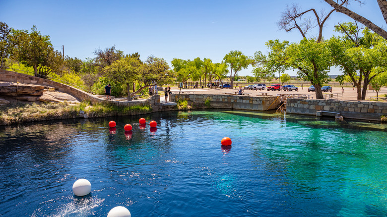 A blue pool of water surrounded by sand colored stones and cars in the distance