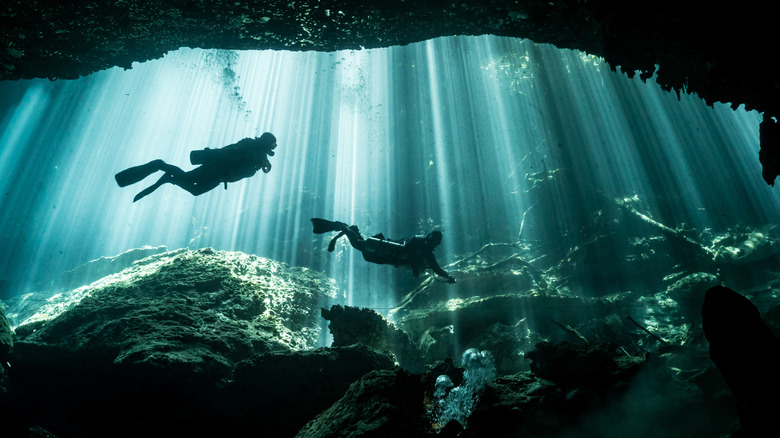 Two divers swim through an opening underwater backlit by rays of sunlight streaming through the water