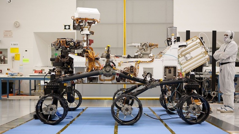 A profile view of a six-wheeled rover in a lab with a person in white clothing standing next to it