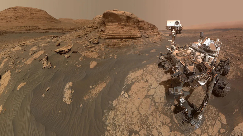 A six-whelled robotic crawler covered in cameras and instruments sits on the red-orange hued terrain of rocks and dust on the Martian surface