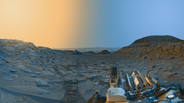 The Curiosity rover sits in the bottom right in a image depicting a panorama of the Martian surface with a reddish orange haze fading left to right into light blue in the sky