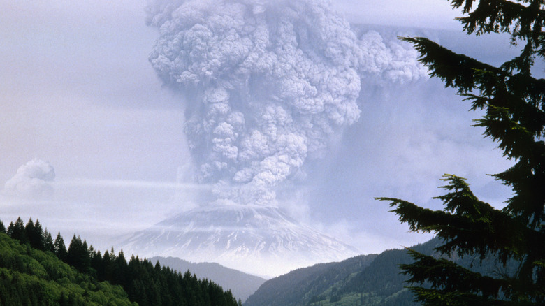 A volcano in the distance spews ash and smoke miles into the atmosphere with green hills and trees in the foreground