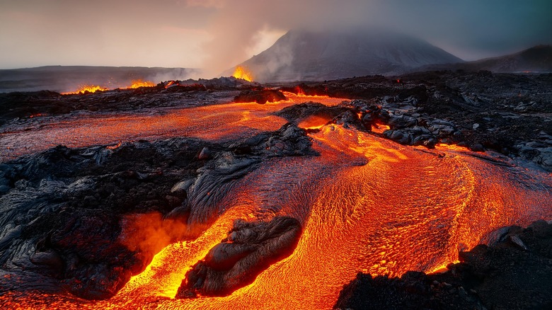 An orange-red lava river flows into the foreground with a volcano peak shrouded in mist and gases in the background