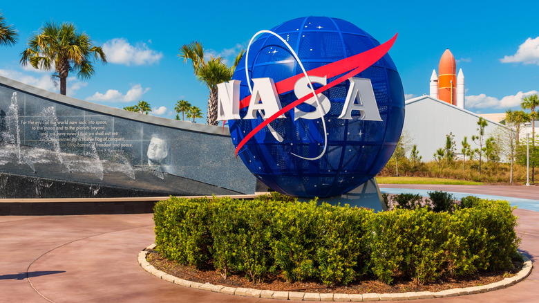 NASA globe logo (center) with Kennedy mural/water feature (left) and orange rocket (right) at the Kennedy Space Center in Florida