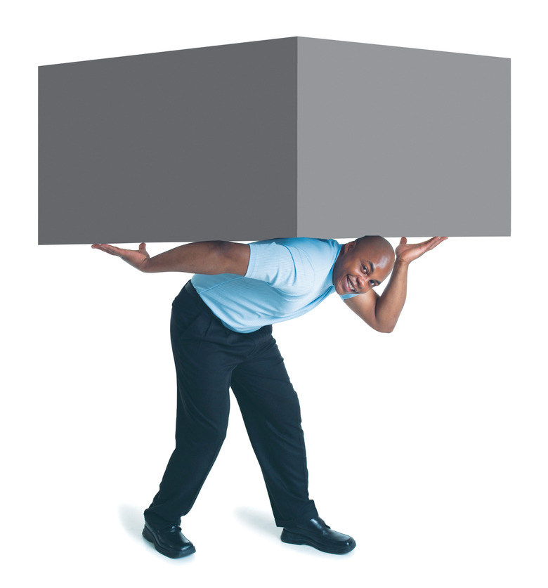 a young african american male in black pants and a blue shirt holds a giant box up with his arms and back