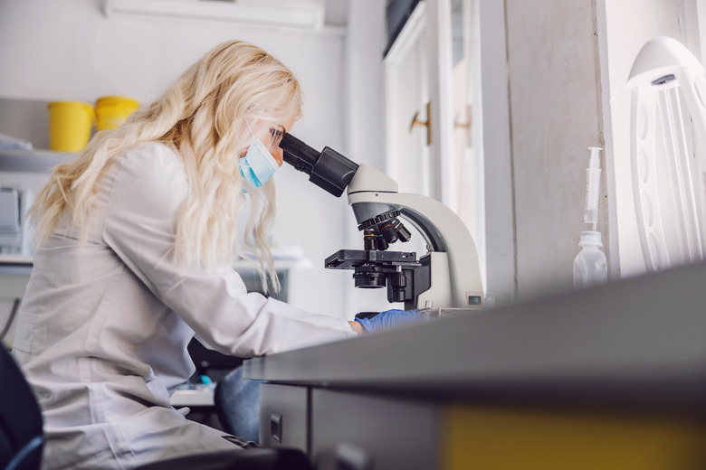 Side view of attractive blond female lab assistant with rubber gloves and face mask sitting in laboratory during corona virus and using microscope.