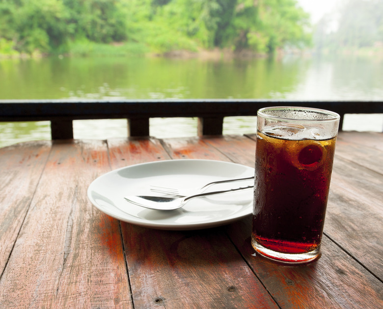 A glass of cola with ice on an outdoor table by a river.