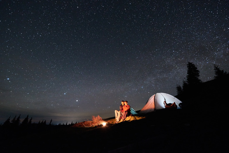 Night camping. Romantic couple tourists have a rest at a campfire near illuminated tent under beautiful night starry sky. Astrophotography