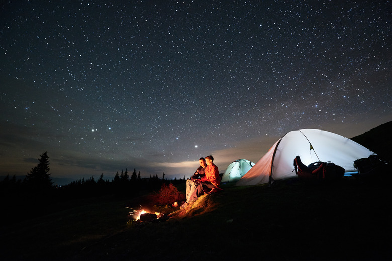 Night camping in the mountains. Man and woman tourists sitting at a campfire near two illuminated tents under starry sky at night. Long exposure