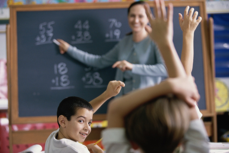 Female teacher pointing to a chalkboard in a classroom with her students raising their hands