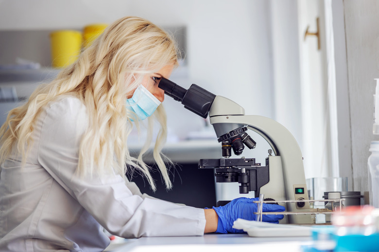 Side view of attractive blond female lab assistant with rubber gloves and face mask sitting in laboratory during corona virus and using microscope.