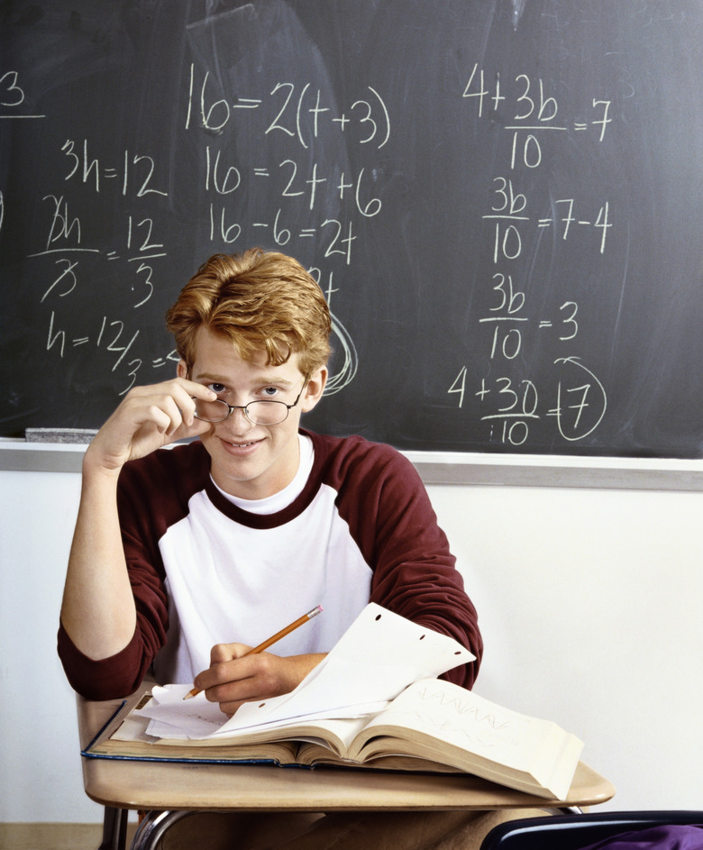 An Intelligent Looking Student Working at a Desk in a Classroom, with a Blackboard Behind him Covered in Algebra Equations