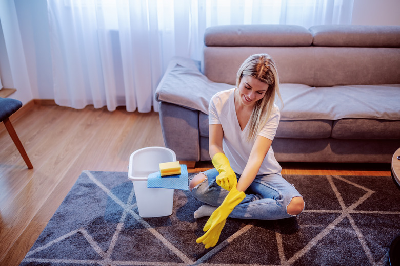 Smiling worthy caucasian blond young housewife sitting on carpet in living room with legs crossed and putting on rubber gloved. Next to her is bucket. Housework.