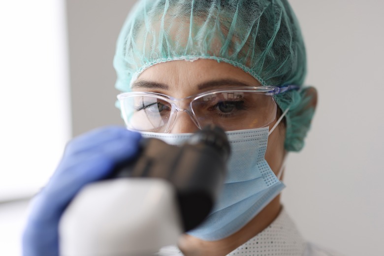 Researcher wearing protective mask and gloves looks through microscope.