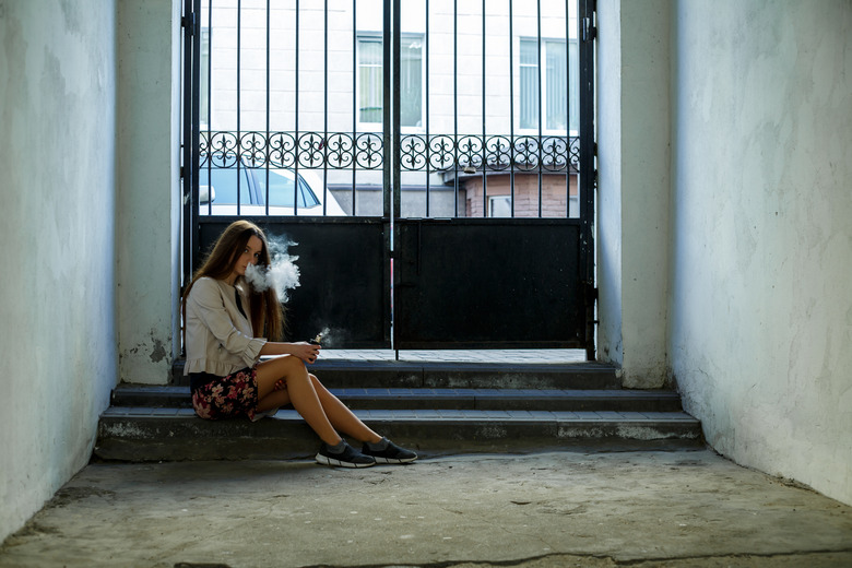 Vape teenager. Young cute girl in casual clothes smokes an electronic cigarette near the wall in prison cell. Bad habit.